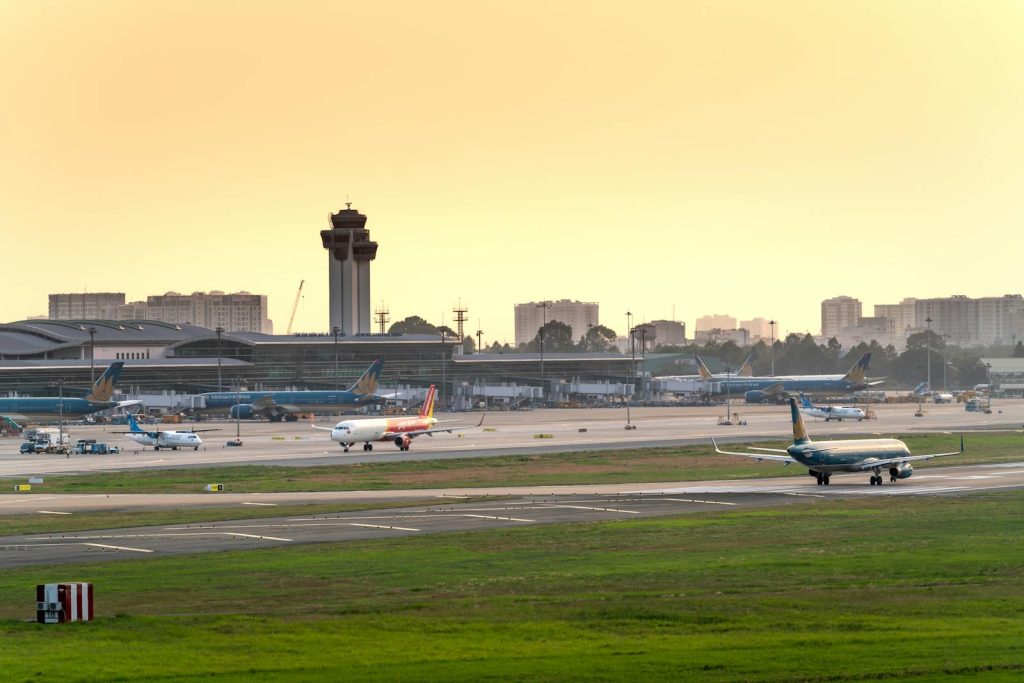 Airplanes prepare for departure on a busy airport runway at sunset, with control tower and city skyline visible.